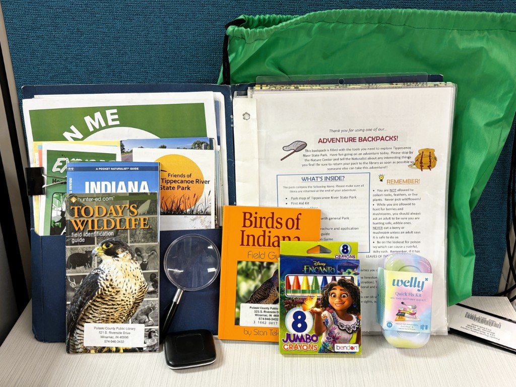 a variety of items in front of green drawstring backpack, including folder containing educational leaflets and brochures ; "Birds of Indiana" field guide book ; a first aid kit ; Barlow 65" measuring tape ; small magnifying glass ; 8 jumbo crayons