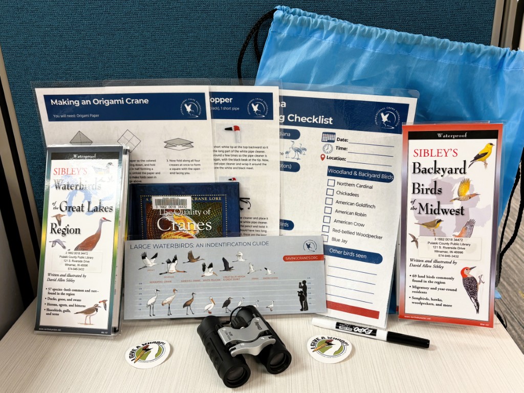 a variety of items in front of a blue drawstring backpack, including Sibley’s Waterbirds of the Great Lakes and Backyard Birds of the Midwest guides; other guides; "The Quality of Cranes" small book; A birding checklist and marker; Binoculars; Whooping Crane stickers; Crane craft instructions