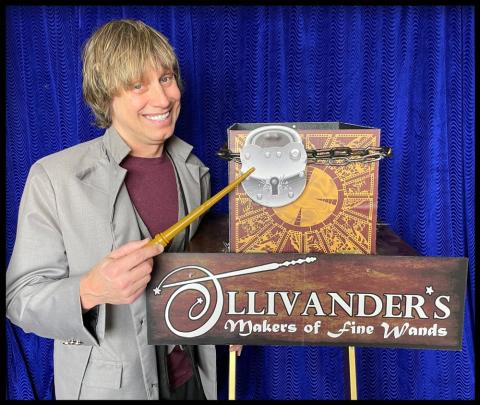 magician Rusty Ammerman holding a wand next to an oversized lock and a sign that says "Ollivander's"