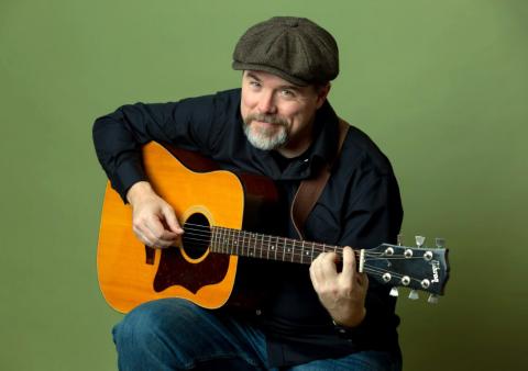 a man with a goatee wearing a flat cap holding a guitar and slightly smiling