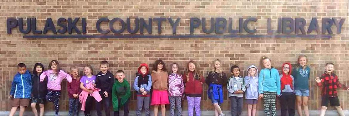 Young children standing in a row outside underneath the "Pulaski County Public Library" sign on the front of the library building.
