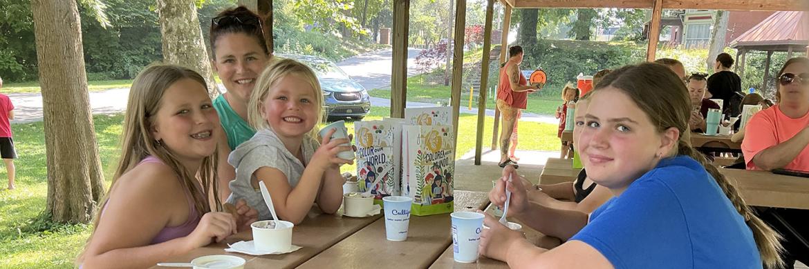 A family smiling and enjoying ice cream at a picnic table in the pavilion next to the Artesian Well at the library's 2025 Summer Reading Party.