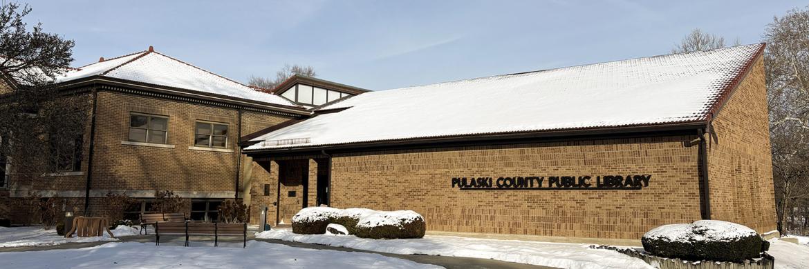 Exterior the Pulaski County Public Library building in winter, with snow on the roof, bushes, and grass. On the front of the tan brick building are a single row of black metal letters that say "Pulaski County Public Library."